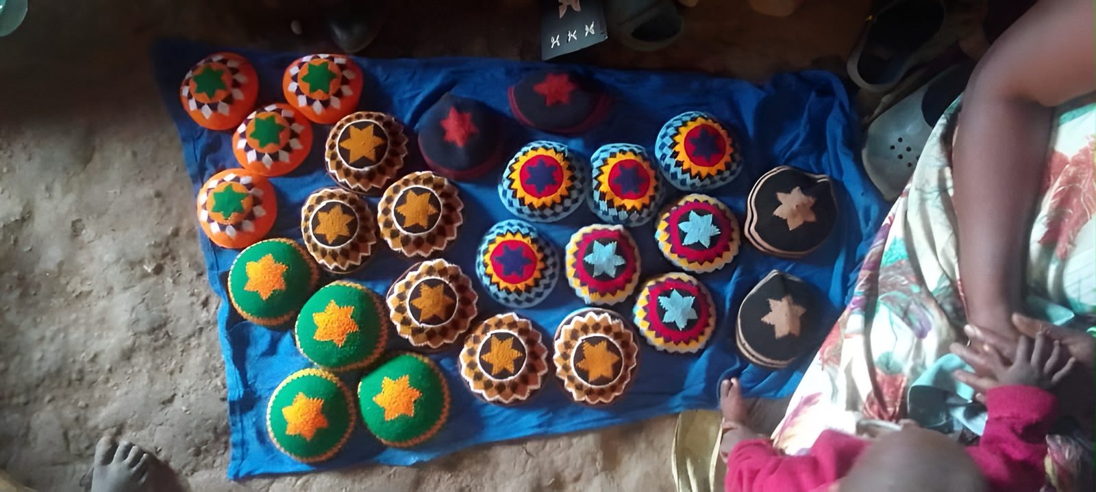 Two dozen crocheted kippot, each with a Magen David at the crown, laid out on a blue cloth on the floor of a home. A mother sits beside them with her baby.