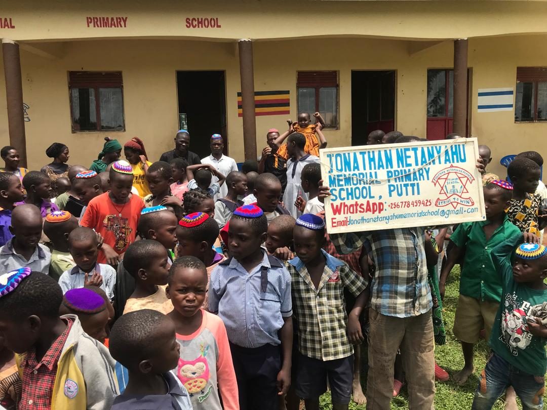 Students in crocheted kippot crowd in front of the school, behind a hand-painted sign reading Jonathan Netanyahu Memorial Primary School, Putti.