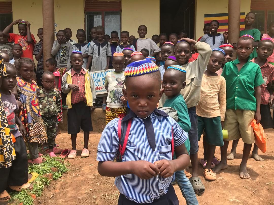A young student in a striped school uniform and a crocheted kippah stands in front of his classmates outside the school.